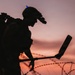A silhouetted military or security personnel figure is photographed against a vivid orange-pink sunset sky. The individual is equipped with a tactical helmet fitted with night-vision goggles and a blue indicator light, and is holding what appears to be a metal detector or mine-detection device. Coils of razor/concertina wire are visible in the foreground, suggesting a secured perimeter or demining operation.
