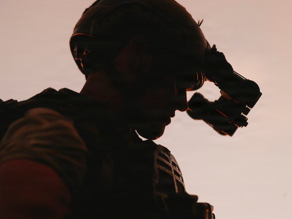 A silhouetted figure in tactical military gear is photographed in dramatic low-light conditions against a pale, warm-toned sky. The subject appears to be adjusting or deploying night vision goggles mounted on a combat helmet. The image is heavily backlit, creating a strong silhouette effect with warm orange rim lighting outlining the figure's profile.