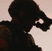 A silhouetted figure in tactical military gear is photographed in dramatic low-light conditions against a pale, warm-toned sky. The subject appears to be adjusting or deploying night vision goggles mounted on a combat helmet. The image is heavily backlit, creating a strong silhouette effect with warm orange rim lighting outlining the figure's profile.