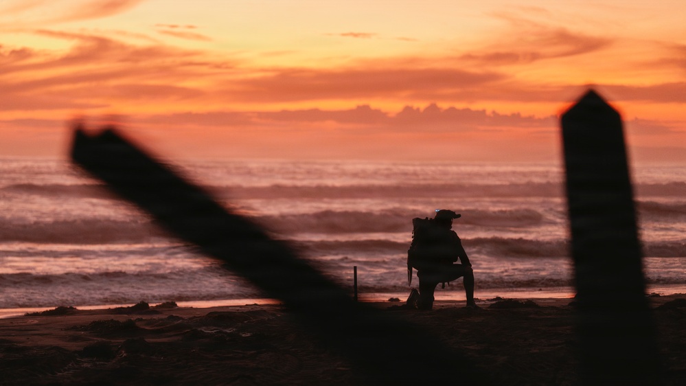A silhouetted figure wearing a backpack and helmet crouches on a beach at sunset or sunrise, with vivid orange and red skies reflected over ocean waves. Large diagonal wooden posts or pilings are visible in the foreground, blurred due to shallow depth of field. The composition creates a dramatic, moody atmosphere with the lone figure framed against the colorful horizon.