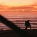 A silhouetted figure wearing a backpack and helmet crouches on a beach at sunset or sunrise, with vivid orange and red skies reflected over ocean waves. Large diagonal wooden posts or pilings are visible in the foreground, blurred due to shallow depth of field. The composition creates a dramatic, moody atmosphere with the lone figure framed against the colorful horizon.