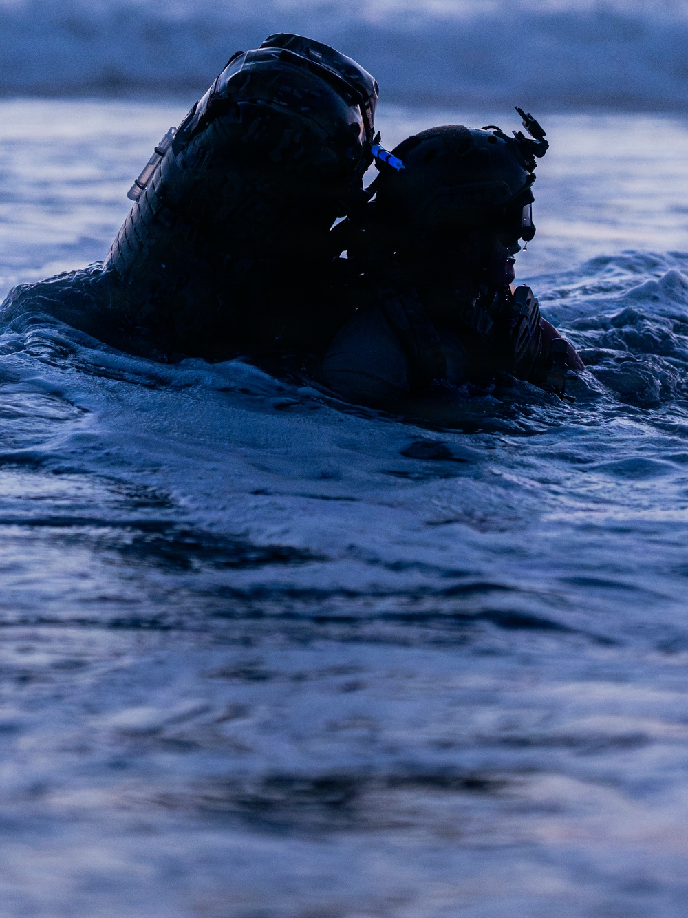 Two heavily equipped individuals are partially submerged in open water during low-light conditions, likely dusk or dawn. Both are wearing tactical helmets with mounted equipment, suggesting a military or special operations context. The image is taken at water level, emphasizing the aquatic environment and the silhouetted figures.