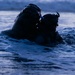 Two heavily equipped individuals are partially submerged in open water during low-light conditions, likely dusk or dawn. Both are wearing tactical helmets with mounted equipment, suggesting a military or special operations context. The image is taken at water level, emphasizing the aquatic environment and the silhouetted figures.