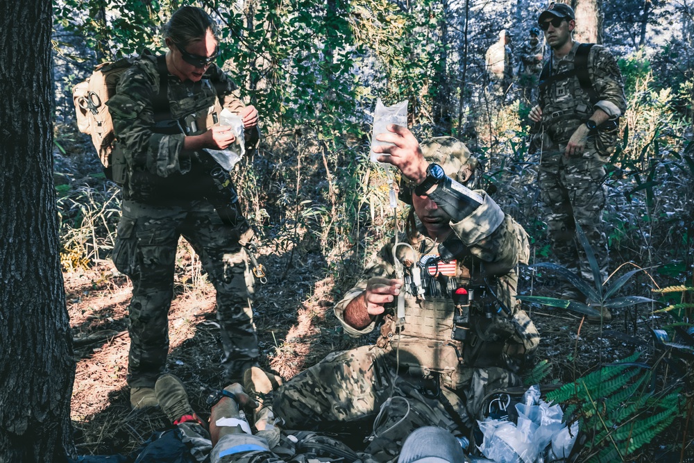Military personnel in camouflage uniforms are conducting a tactical combat casualty care (TCCC) exercise in a wooded outdoor environment. Two medics are actively treating a simulated casualty on the ground, with one holding up what appears to be an IV fluid bag. A third uniformed individual stands in the background observing the scenario.