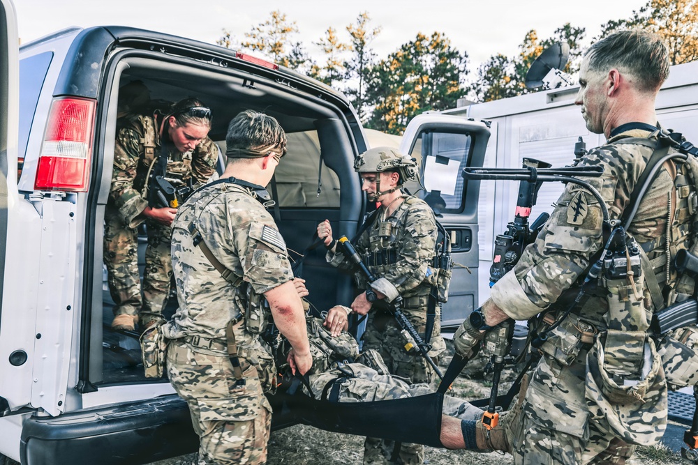 A group of military personnel in camouflage uniforms are loading or unloading a casualty on a stretcher into the back of a white van. The scene appears to depict a tactical medical evacuation exercise or operation, with multiple soldiers actively participating in the patient transfer. The setting is an outdoor staging area with trees and additional vehicles visible in the background.