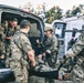 A group of military personnel in camouflage uniforms are loading or unloading a casualty on a stretcher into the back of a white van. The scene appears to depict a tactical medical evacuation exercise or operation, with multiple soldiers actively participating in the patient transfer. The setting is an outdoor staging area with trees and additional vehicles visible in the background.
