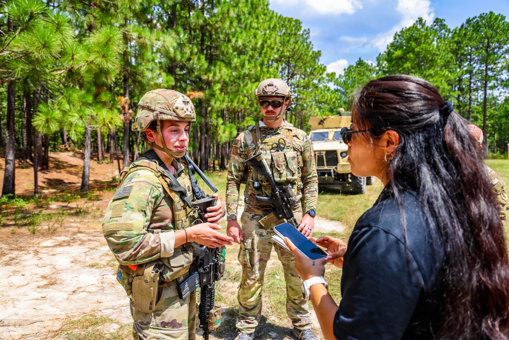 Two soldiers in full combat gear and camouflage uniforms are engaged in conversation with a civilian woman holding a tablet/phone in an outdoor training area. The setting appears to be a military training ground surrounded by pine trees, with a military tactical vehicle visible in the background. The interaction suggests a briefing, interview, or demonstration scenario.