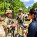 Two soldiers in full combat gear and camouflage uniforms are engaged in conversation with a civilian woman holding a tablet/phone in an outdoor training area. The setting appears to be a military training ground surrounded by pine trees, with a military tactical vehicle visible in the background. The interaction suggests a briefing, interview, or demonstration scenario.