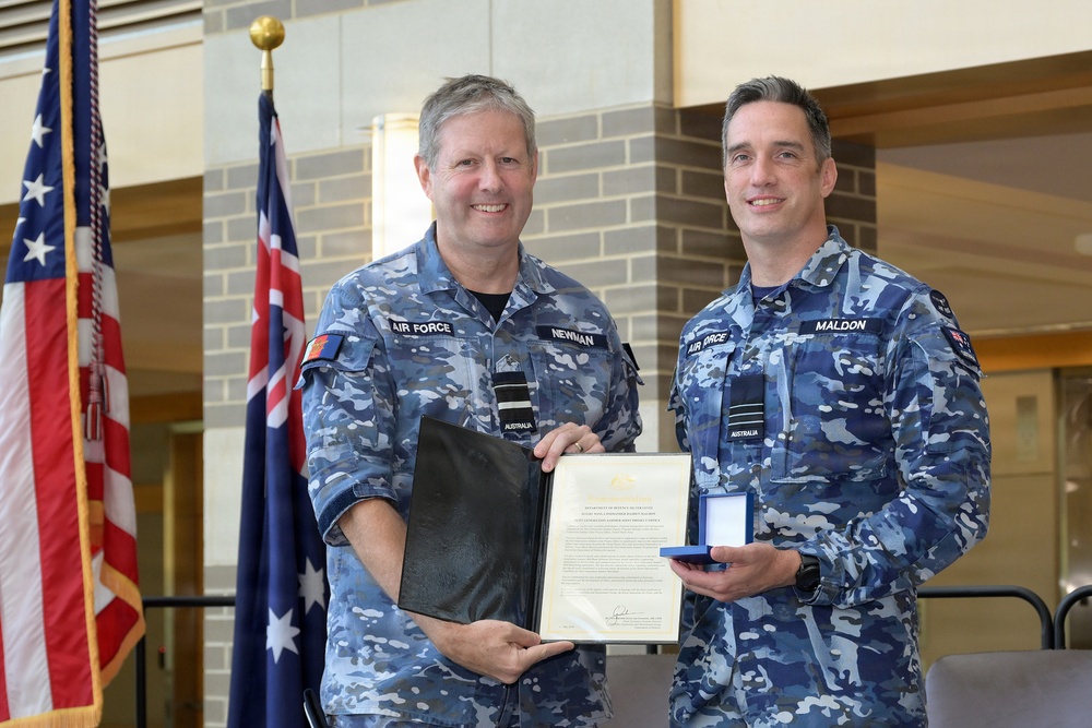 Two Royal Australian Air Force personnel in blue multicam uniforms are posing together during what appears to be a formal award or commendation ceremony. The individual on the left, identified by name tape as 'NEWMAN,' and the individual on the right, identified as 'MALDON,' are jointly holding a framed certificate and a medal in a presentation box. An Australian flag and a United States flag are visible in the background, suggesting a joint military event.