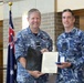 Two Royal Australian Air Force personnel in blue multicam uniforms are posing together during what appears to be a formal award or commendation ceremony. The individual on the left, identified by name tape as 'NEWMAN,' and the individual on the right, identified as 'MALDON,' are jointly holding a framed certificate and a medal in a presentation box. An Australian flag and a United States flag are visible in the background, suggesting a joint military event.
