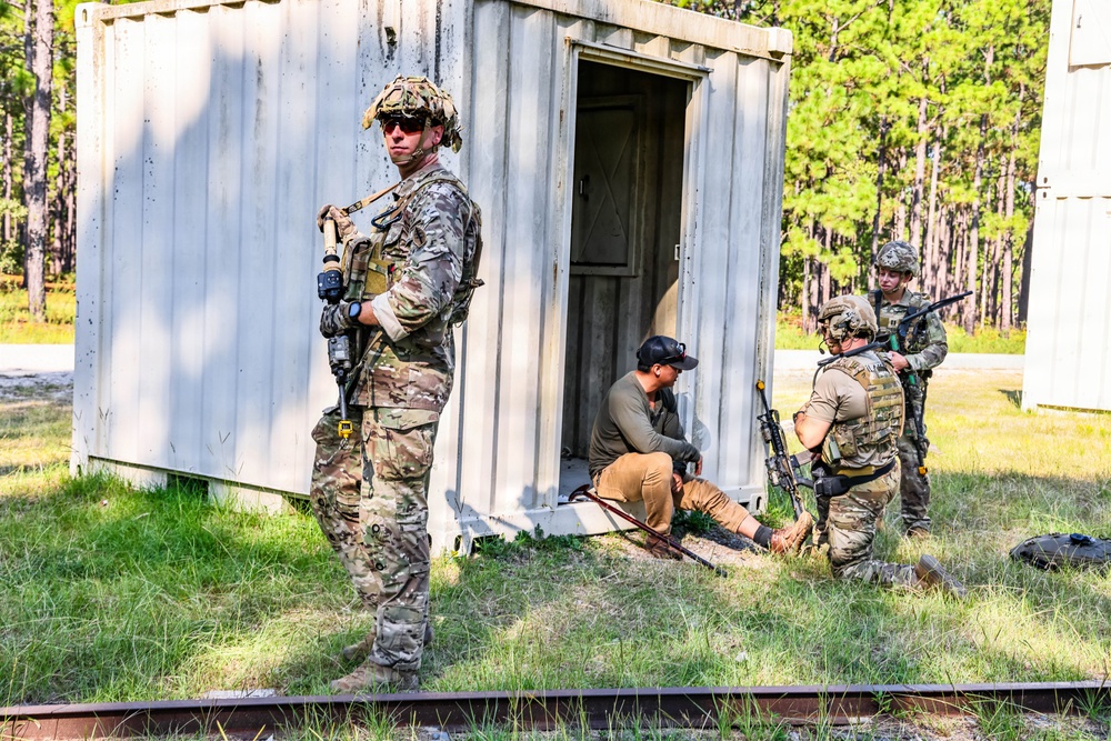 The image depicts what appears to be a military training exercise at an outdoor facility surrounded by pine trees. Several soldiers in multicam/OCP camouflage uniforms with tactical gear are interacting with a civilian role-player near a small metal storage container structure. The scenario appears to simulate a detainee handling or personnel search procedure.