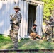 The image depicts what appears to be a military training exercise at an outdoor facility surrounded by pine trees. Several soldiers in multicam/OCP camouflage uniforms with tactical gear are interacting with a civilian role-player near a small metal storage container structure. The scenario appears to simulate a detainee handling or personnel search procedure.