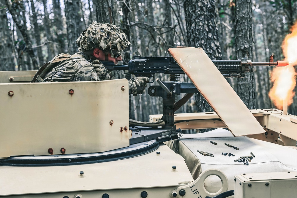 A soldier in camouflage uniform and helmet is firing a machine gun from the turret of a military vehicle in a forested environment. A muzzle flash is clearly visible at the end of the barrel, and spent brass casings are scattered on the vehicle's surface. The vehicle appears to be a light armored or wheeled military platform painted in a tan/desert color scheme.