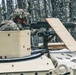 A soldier in camouflage uniform and helmet is firing a machine gun from the turret of a military vehicle in a forested environment. A muzzle flash is clearly visible at the end of the barrel, and spent brass casings are scattered on the vehicle's surface. The vehicle appears to be a light armored or wheeled military platform painted in a tan/desert color scheme.