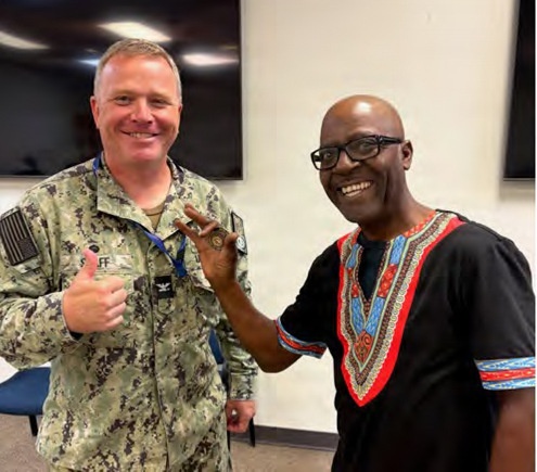 Two men are posing together for a photo in what appears to be an indoor facility, both smiling broadly. One man is wearing a U.S. Navy digital camouflage uniform with a name tag and rank insignia, while the other is wearing a black dashiki-style shirt with colorful embroidery. Both appear to be in good spirits, making hand gestures toward the camera.