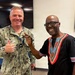 Two men are posing together for a photo in what appears to be an indoor facility, both smiling broadly. One man is wearing a U.S. Navy digital camouflage uniform with a name tag and rank insignia, while the other is wearing a black dashiki-style shirt with colorful embroidery. Both appear to be in good spirits, making hand gestures toward the camera.