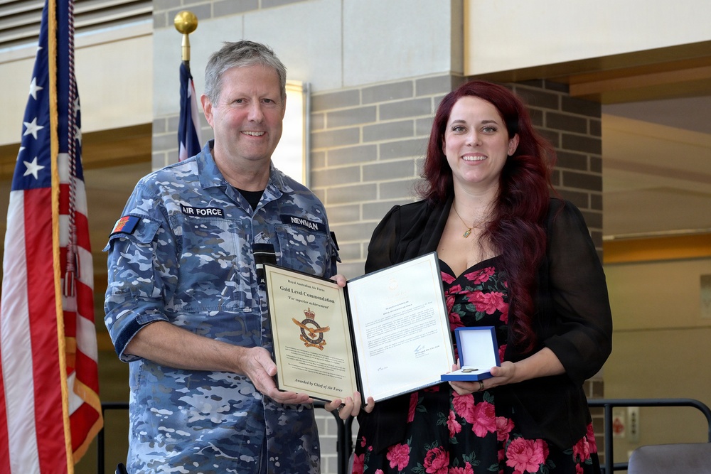 A Royal Australian Air Force officer in camouflage uniform is presenting a Gold Level Commendation award to a civilian woman with red hair. The ceremony appears to take place in an indoor facility with flags visible in the background. Both individuals are smiling and jointly holding the award documents and medal case.