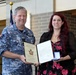 A Royal Australian Air Force officer in camouflage uniform is presenting a Gold Level Commendation award to a civilian woman with red hair. The ceremony appears to take place in an indoor facility with flags visible in the background. Both individuals are smiling and jointly holding the award documents and medal case.