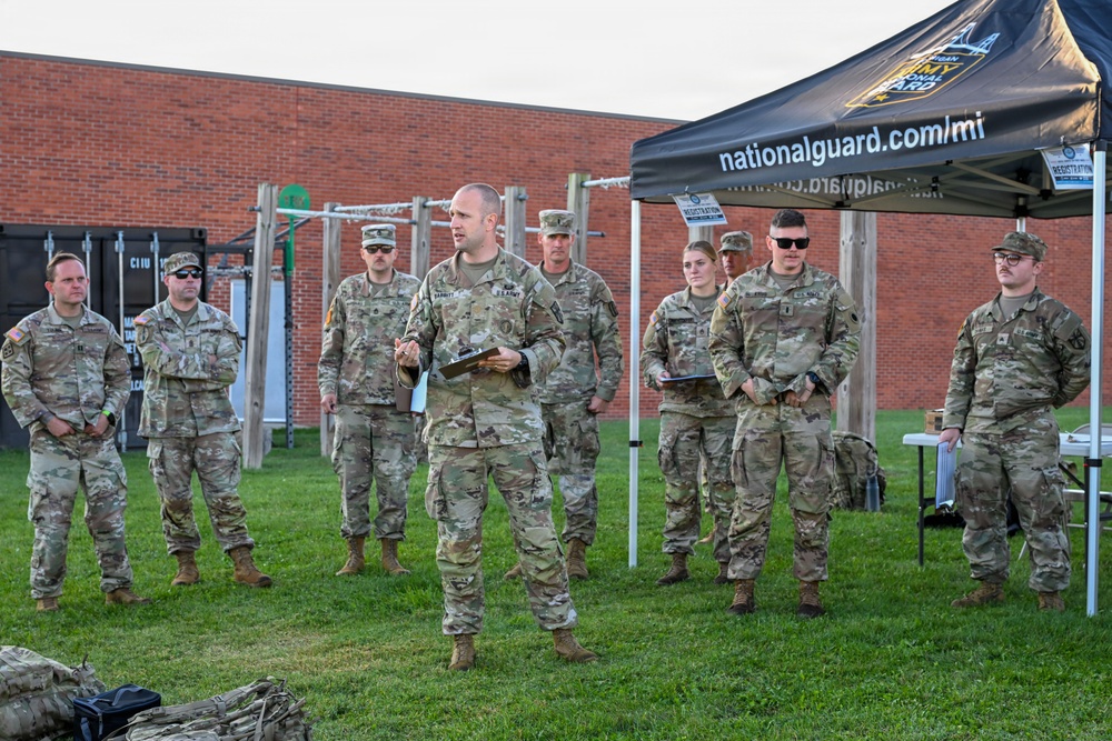 A group of U.S. Army National Guard soldiers are assembled outdoors on a grass field, with one soldier in the center appearing to address the group while holding a clipboard. A black canopy tent bearing the text 'nationalguard.com/mi' and the Michigan Army National Guard logo is visible to the right. The setting appears to be a military training or recruitment event at a facility with a brick building and outdoor fitness equipment in the background.
