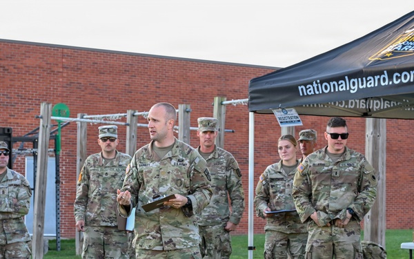A group of U.S. Army National Guard soldiers are assembled outdoors on a grass field, with one soldier in the center appearing to address the group while holding a clipboard. A black canopy tent bearing the text 'nationalguard.com/mi' and the Michigan Army National Guard logo is visible to the right. The setting appears to be a military training or recruitment event at a facility with a brick building and outdoor fitness equipment in the background.