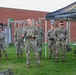 A group of U.S. Army National Guard soldiers are assembled outdoors on a grass field, with one soldier in the center appearing to address the group while holding a clipboard. A black canopy tent bearing the text 'nationalguard.com/mi' and the Michigan Army National Guard logo is visible to the right. The setting appears to be a military training or recruitment event at a facility with a brick building and outdoor fitness equipment in the background.