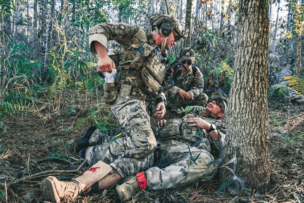 The image depicts what appears to be a military tactical combat casualty care (TCCC) training exercise or real field medical response in a wooded environment. Two standing/crouching soldiers in multicam uniforms are attending to a downed soldier lying on the ground near a tree. The casualty has visible simulated or real blood on the lower leg and a red tourniquet applied, indicating a leg wound scenario.