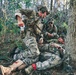 The image depicts what appears to be a military tactical combat casualty care (TCCC) training exercise or real field medical response in a wooded environment. Two standing/crouching soldiers in multicam uniforms are attending to a downed soldier lying on the ground near a tree. The casualty has visible simulated or real blood on the lower leg and a red tourniquet applied, indicating a leg wound scenario.