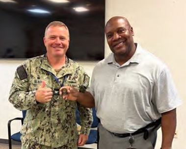 Two men are posing together indoors, both smiling at the camera. The man on the left is wearing a U.S. Navy NWU Type III (digital camouflage) uniform with a name tape reading 'SLAF-' (partially visible) and is giving a thumbs up while holding what appears to be a challenge coin. The man on the right is in civilian attire (gray polo shirt) and is also holding the coin jointly.
