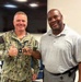 Two men are posing together indoors, both smiling at the camera. The man on the left is wearing a U.S. Navy NWU Type III (digital camouflage) uniform with a name tape reading 'SLAF-' (partially visible) and is giving a thumbs up while holding what appears to be a challenge coin. The man on the right is in civilian attire (gray polo shirt) and is also holding the coin jointly.