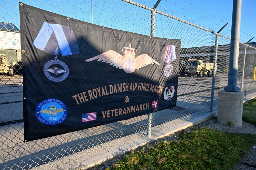 A large black banner is attached to a chain-link fence topped with barbed wire, advertising 'The Royal Danish Air Force March &amp; Veteranmarch.' The banner features military insignia, medals, wings with a crown, and both American and Danish flags, along with a '2026' patch labeled 'Flyver March / Danish Air Force March.' The setting appears to be a military installation or base, with military vehicles and buildings visible in the background.