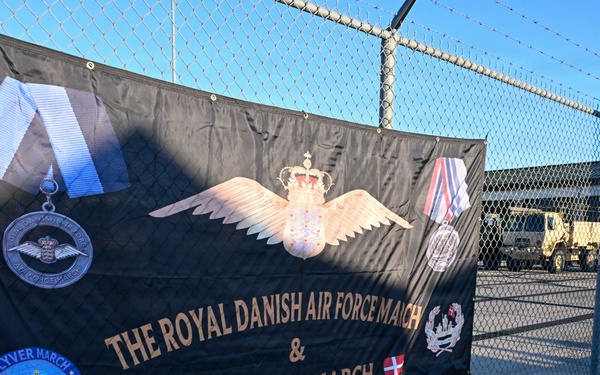 A large black banner is attached to a chain-link fence topped with barbed wire, advertising 'The Royal Danish Air Force March &amp; Veteranmarch.' The banner features military insignia, medals, wings with a crown, and both American and Danish flags, along with a '2026' patch labeled 'Flyver March / Danish Air Force March.' The setting appears to be a military installation or base, with military vehicles and buildings visible in the background.