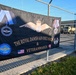 A large black banner is attached to a chain-link fence topped with barbed wire, advertising 'The Royal Danish Air Force March &amp; Veteranmarch.' The banner features military insignia, medals, wings with a crown, and both American and Danish flags, along with a '2026' patch labeled 'Flyver March / Danish Air Force March.' The setting appears to be a military installation or base, with military vehicles and buildings visible in the background.