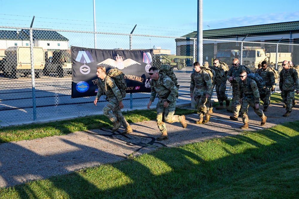 A group of military personnel in camouflage uniforms and tactical backpacks are marching along a paved path at what appears to be a military installation. A black banner in the background references an 'Air Force March' event and displays multiple military insignia including what appears to be NATO-affiliated and Danish symbols. The setting includes chain-link fencing, military vehicles, and green-roofed buildings.