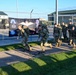 A group of military personnel in camouflage uniforms and tactical backpacks are marching along a paved path at what appears to be a military installation. A black banner in the background references an 'Air Force March' event and displays multiple military insignia including what appears to be NATO-affiliated and Danish symbols. The setting includes chain-link fencing, military vehicles, and green-roofed buildings.