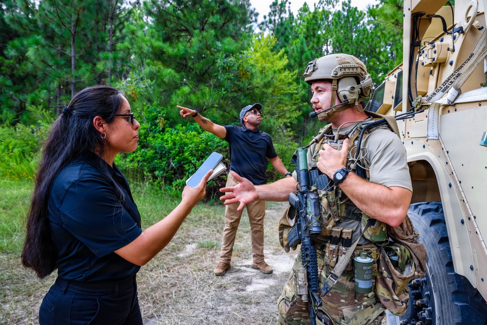 A fully equipped military or tactical operator in multicam camouflage, combat helmet, and body armor is engaged in a conversation with a civilian woman holding a tablet device, near a tan military vehicle. A second civilian man in the background is pointing toward the tree line. The setting appears to be a rural or training area surrounded by pine trees.