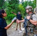 A fully equipped military or tactical operator in multicam camouflage, combat helmet, and body armor is engaged in a conversation with a civilian woman holding a tablet device, near a tan military vehicle. A second civilian man in the background is pointing toward the tree line. The setting appears to be a rural or training area surrounded by pine trees.