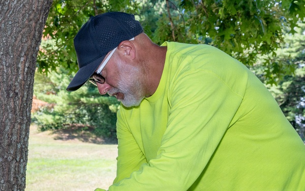 A middle-aged man wearing a neon yellow long-sleeve shirt and black cap is installing or adjusting a green trail camera on a tree trunk. The setting appears to be a park or open wooded area with well-maintained grass in the background. The device mounted on the tree bears a logo consistent with wildlife monitoring equipment.