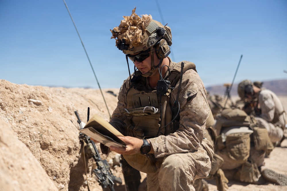 A soldier in full combat gear is seated in a desert trench or fighting position, reading from a notebook or field manual. At least one additional soldier is visible in the background in a crouched position. The environment is arid and open, consistent with a desert training or operational environment.
