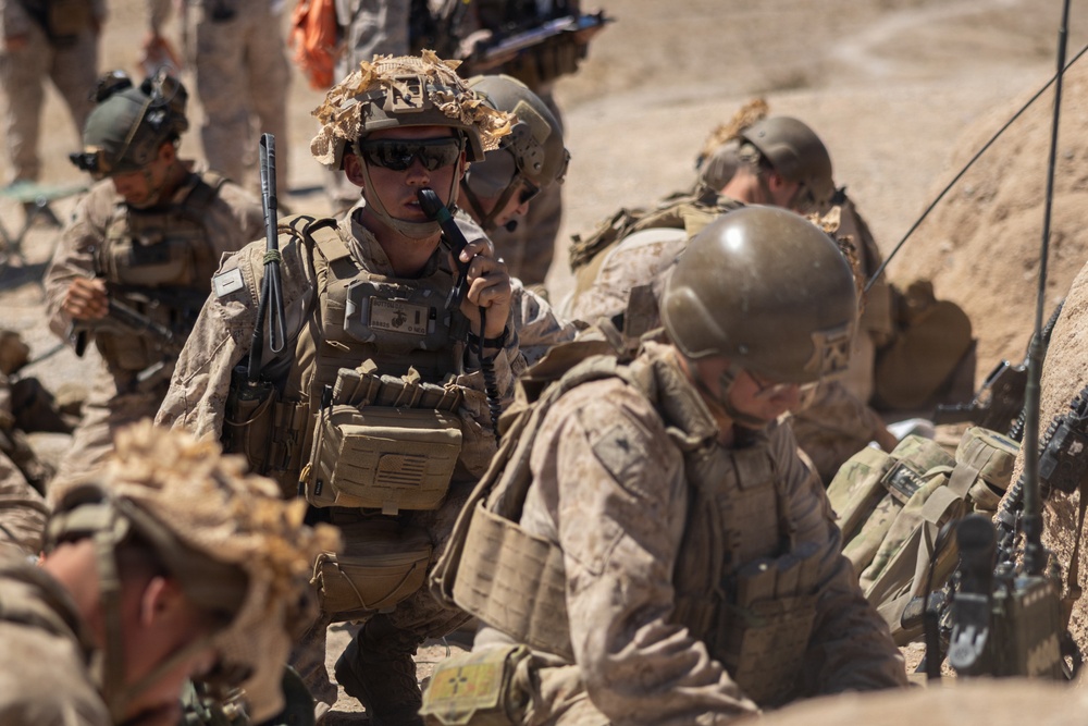 The image shows a group of military personnel in desert camouflage uniforms and tactical gear operating in an arid, sandy environment. One central figure is actively communicating via a handheld radio while others appear to be in a low-profile or crouching position. The scene suggests a field operation, training exercise, or tactical coordination scenario.