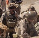 The image shows a group of military personnel in desert camouflage uniforms and tactical gear operating in an arid, sandy environment. One central figure is actively communicating via a handheld radio while others appear to be in a low-profile or crouching position. The scene suggests a field operation, training exercise, or tactical coordination scenario.