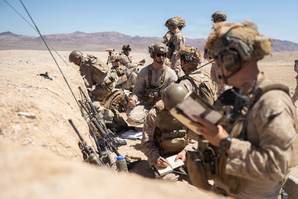 A group of military personnel in desert camouflage uniforms are gathered in a shallow trench or depression in an arid desert environment. They appear to be conducting a briefing or planning session, with some taking notes and others listening. The setting suggests a training exercise or field operation in a desert region.