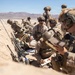 A group of military personnel in desert camouflage uniforms are gathered in a shallow trench or depression in an arid desert environment. They appear to be conducting a briefing or planning session, with some taking notes and others listening. The setting suggests a training exercise or field operation in a desert region.