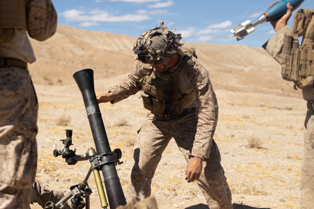 Military personnel in desert camouflage uniforms are operating a mortar system in an arid, desert training environment. The central figure is adjusting or steadying the mortar tube while another individual to the right holds what appears to be a mortar round. The setting and uniforms are consistent with U.S. Marine Corps desert training operations.