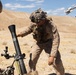 Military personnel in desert camouflage uniforms are operating a mortar system in an arid, desert training environment. The central figure is adjusting or steadying the mortar tube while another individual to the right holds what appears to be a mortar round. The setting and uniforms are consistent with U.S. Marine Corps desert training operations.
