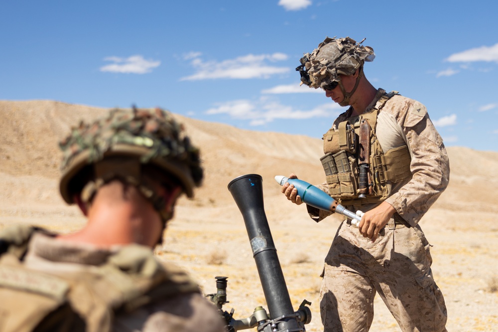 Two military personnel in desert camouflage uniforms are operating a mortar system in an arid, desert training environment. The soldier in the foreground is positioned near the mortar tube while the soldier in the background handles what appears to be a blue-tipped training round. The setting and blue-colored munition strongly suggest a military training exercise rather than a combat operation.