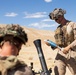 Two military personnel in desert camouflage uniforms are operating a mortar system in an arid, desert training environment. The soldier in the foreground is positioned near the mortar tube while the soldier in the background handles what appears to be a blue-tipped training round. The setting and blue-colored munition strongly suggest a military training exercise rather than a combat operation.