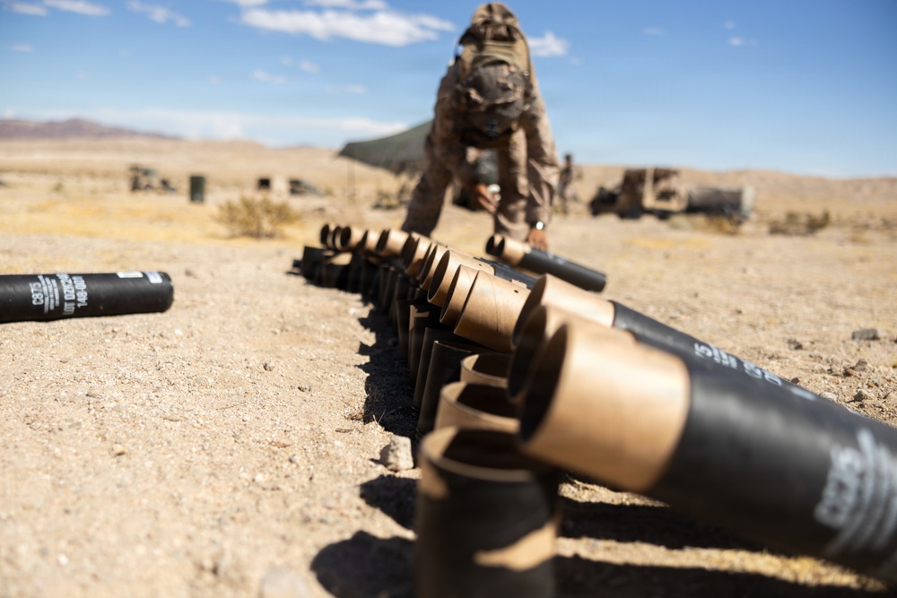 A military service member in desert camouflage uniform is arranging a row of artillery shell casings on sandy desert terrain. The image is taken from a low ground-level perspective, with the shell casings lined up in the foreground and the soldier visible in the background. The setting appears to be a desert training range with military vehicles and equipment visible in the distance.
