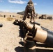 A military service member in desert camouflage uniform is arranging a row of artillery shell casings on sandy desert terrain. The image is taken from a low ground-level perspective, with the shell casings lined up in the foreground and the soldier visible in the background. The setting appears to be a desert training range with military vehicles and equipment visible in the distance.