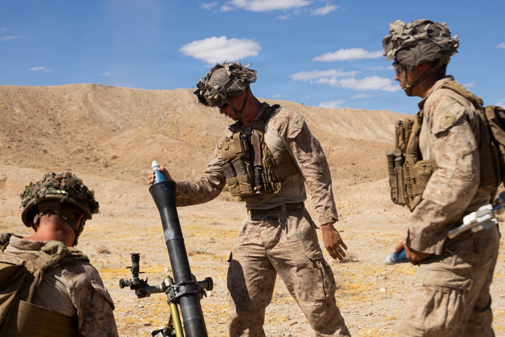 Three military personnel in desert camouflage uniforms are operating a mortar system in an arid, desert training environment. The central figure is handling what appears to be a mortar round above the tube, while a third soldier observes from the right. The setting and equipment suggest a live-fire training exercise.