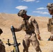 Three military personnel in desert camouflage uniforms are operating a mortar system in an arid, desert training environment. The central figure is handling what appears to be a mortar round above the tube, while a third soldier observes from the right. The setting and equipment suggest a live-fire training exercise.